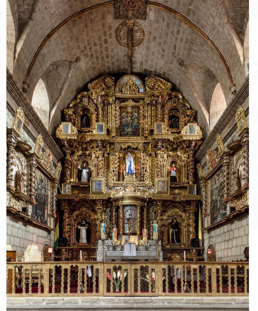 Elaborate gold altar in a church interior, featuring religious art and statues, stone archways and walls, and flowers.