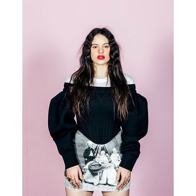 A woman with long dark hair, red lipstick, wearing a black and white top and printed skirt, stands against a light pink background.