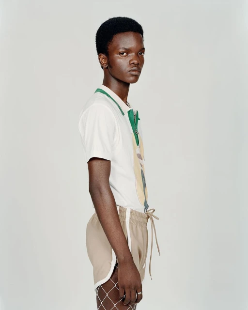 Profile shot of a young Black man in a polo shirt and shorts, posed against a neutral background in a brightly lit studio setting.