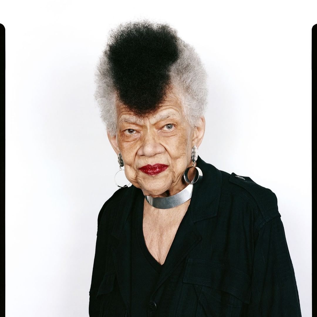 Elderly woman with a distinctive black and grey mohawk, wearing a black jacket and silver accessories in a studio portrait.