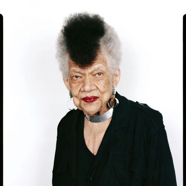 Elderly woman with a distinctive black and grey mohawk, wearing a black jacket and silver accessories in a studio portrait.