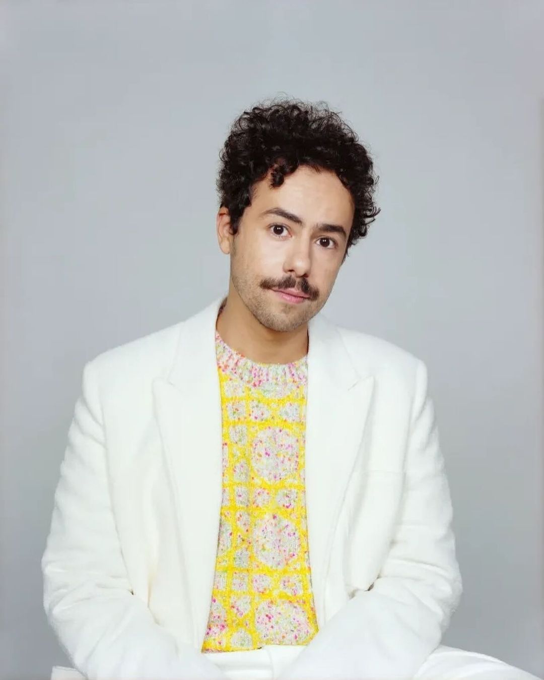 Portrait of a man with curly dark hair and a mustache wearing a white blazer over a patterned yellow sweater against a grey background.