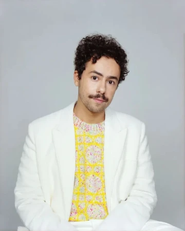 Portrait of a man with curly dark hair and a mustache wearing a white blazer over a patterned yellow sweater against a grey background.
