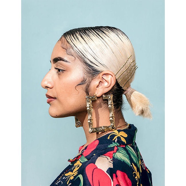Profile portrait of a woman with blonde hair wearing large square gold bamboo earrings and a floral shirt in front of a blue background.