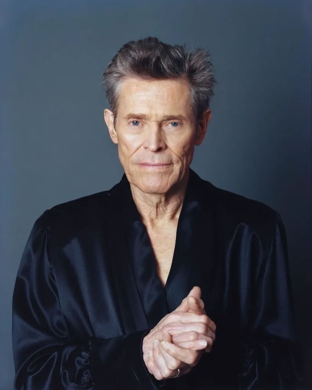 Headshot of Willem Dafoe in a dark robe against a gray backdrop, close-up with hands clasped, neutral expression, and soft studio lighting.