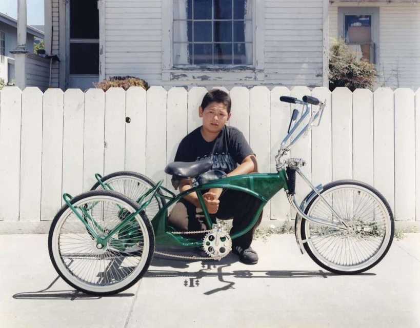 Boy poses with a customized green lowrider tricycle on the sidewalk in front of a white picket fence and a weathered house.