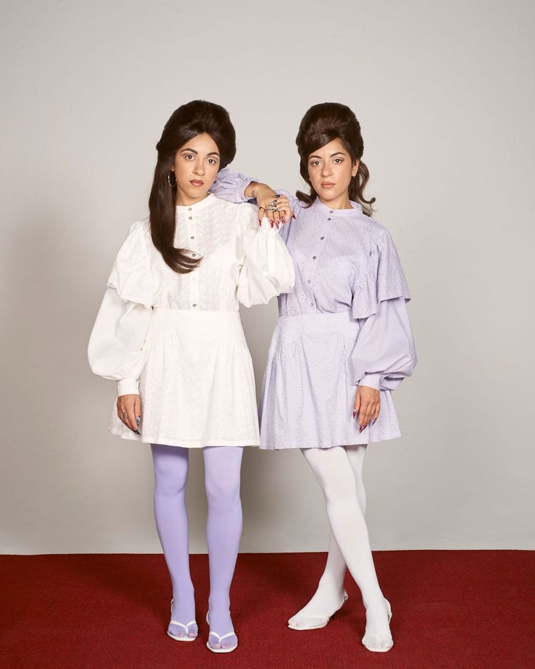 Two women in vintage-inspired dresses with bouffant hairstyles pose in a studio against a neutral backdrop, one with lilac tights and the other with white.