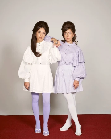 Two women in vintage-inspired dresses with bouffant hairstyles pose in a studio against a neutral backdrop, one with lilac tights and the other with white.