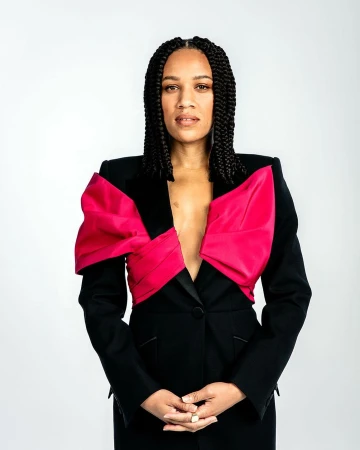 Woman with braided hair wearing a black jacket with vibrant pink lapels against a plain white background
