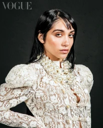 Portrait of a woman wearing a white lace high-neck top and a chunky multi-strand pearl necklace against a dark textured background