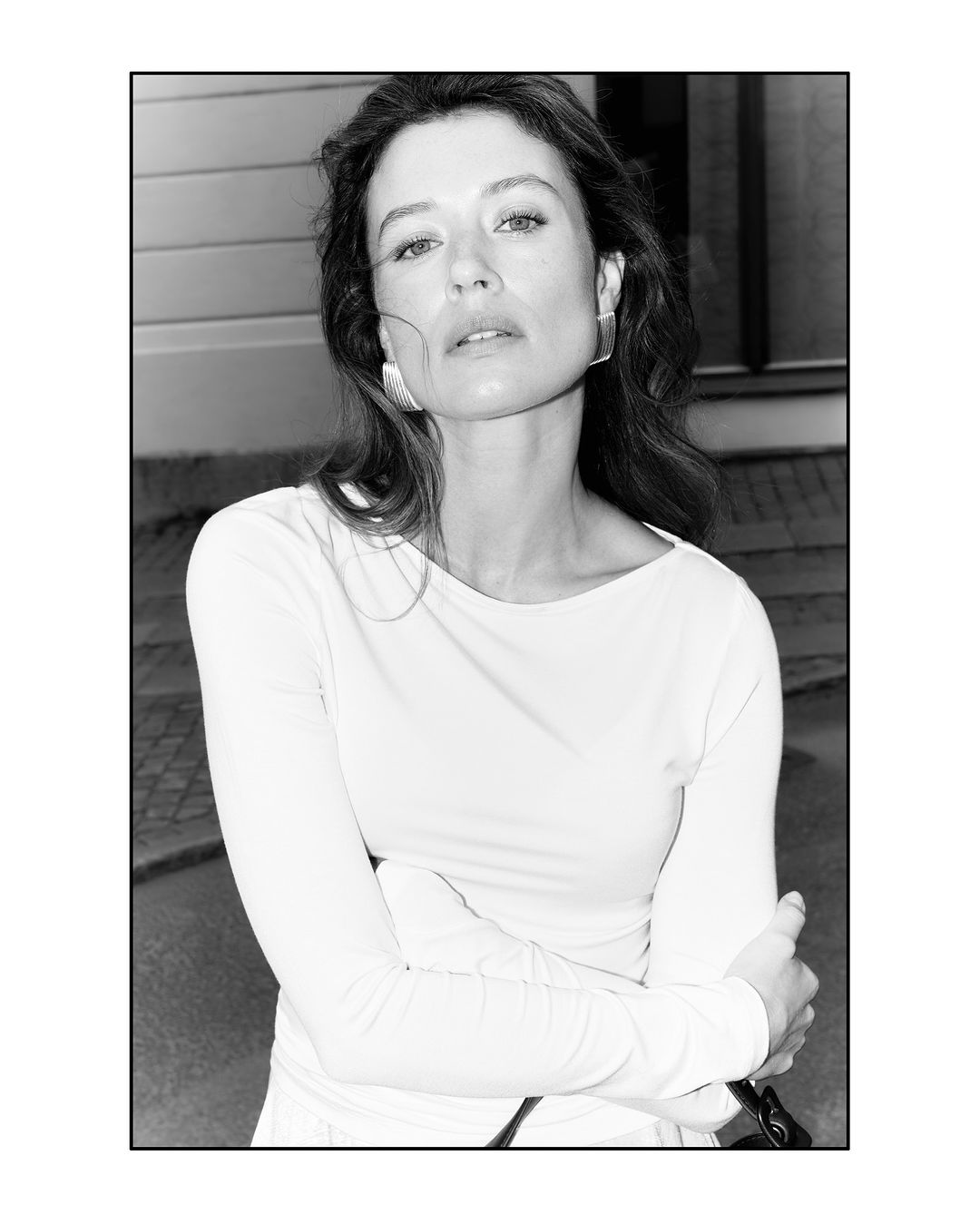 A portrait shot of a fair-skinned woman with wavy brown hair, wearing a white long-sleeved shirt and geometric earrings in an outdoor setting.