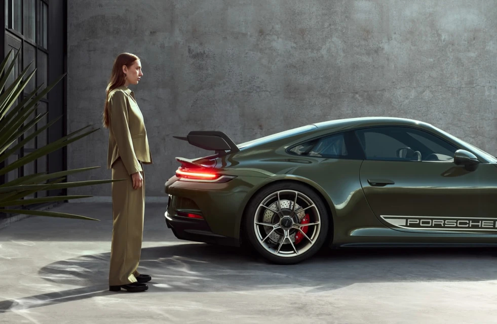 Olive-green Porsche with tail wing parked in front of industrial-style building with woman in matching suit standing alongside.