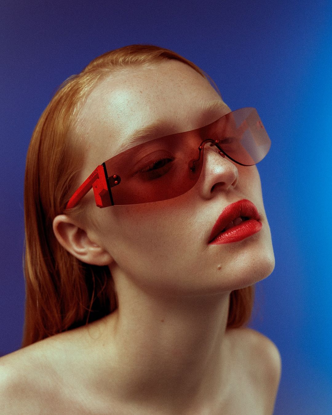 Close-up portrait of a redhead woman with red lipstick and sunglasses on a blue gradient background, demonstrating beauty and fashion.