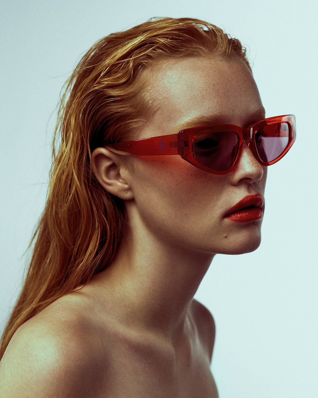 A close-up studio portrait of a red-haired, fair-skinned adult woman wearing translucent red sunglasses and bright red lipstick.