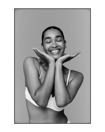 Portrait of a smiling young woman with her eyes closed, holding her hands to her cheeks against a plain background.