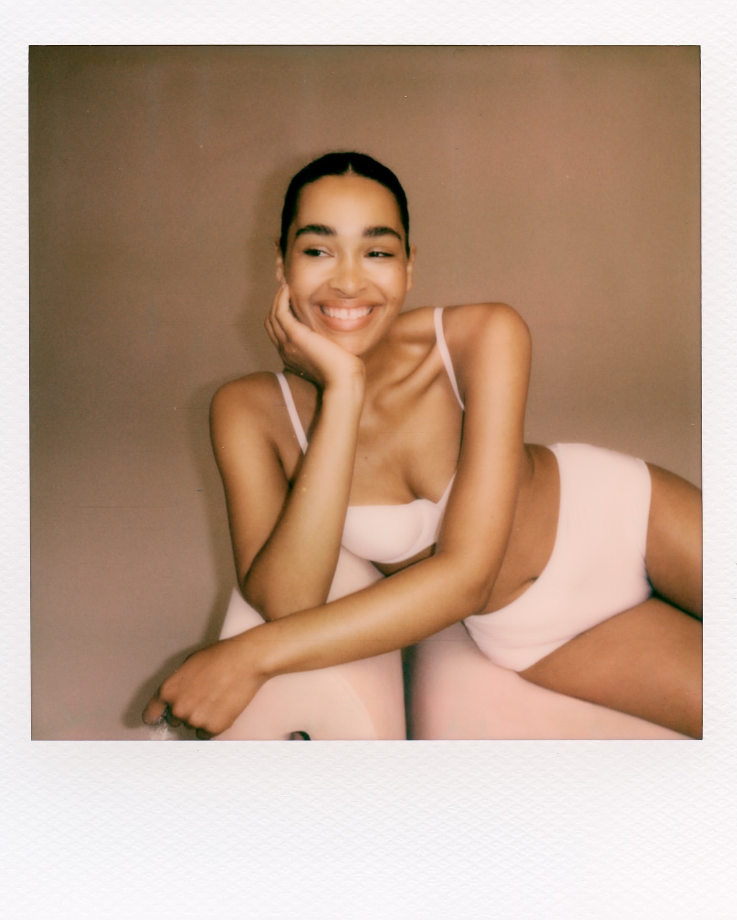 Smiling woman with dark hair and fair skin in a bralette and high-waisted underwear set, reclining against a round object in a photo studio.