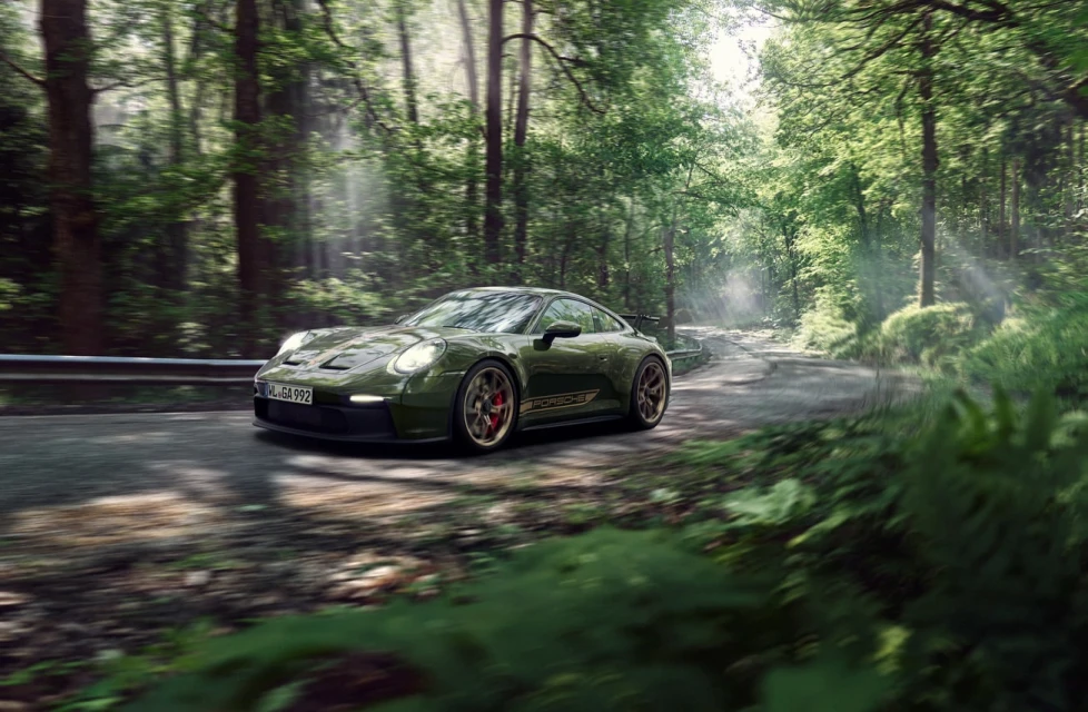 An olive-green Porsche 911 GT3 car drives on an asphalt road through a bright, sunlit forest with green foliage and tall trees.