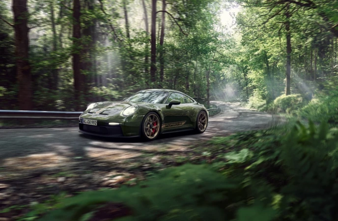 An olive-green Porsche 911 GT3 car drives on an asphalt road through a bright, sunlit forest with green foliage and tall trees.