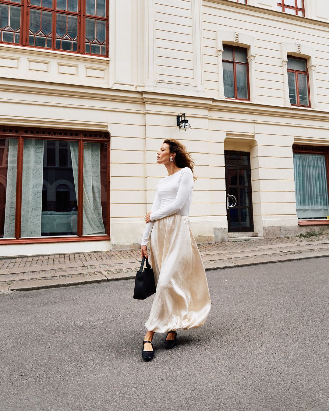 A woman in a white long-sleeve shirt and a satin skirt carries a bag while standing on a city street near a light-beige building.
