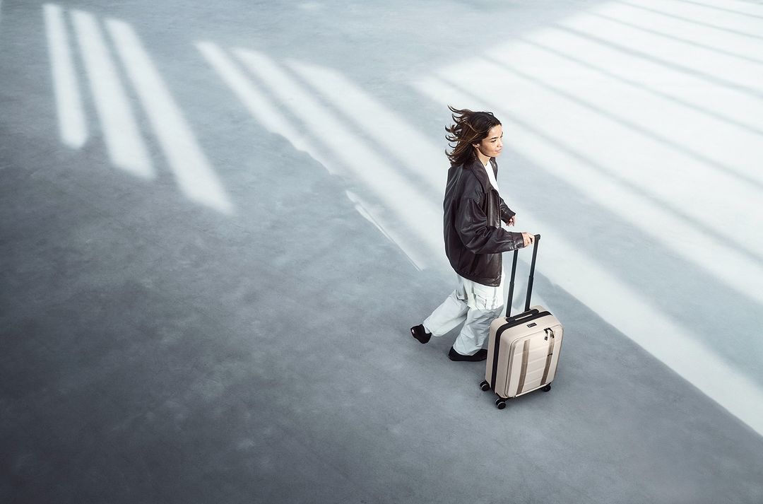 A woman walks in a bright, modern space, pulling a beige rolling suitcase behind her. Light streams through unseen windows above.
