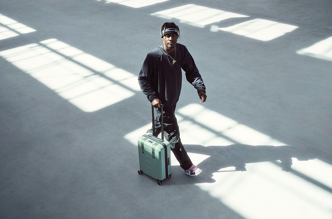 Adult male pulling a suitcase in an airy, open indoor setting with bright sunlight, wearing a black sweatshirt, pants, and headphones.
