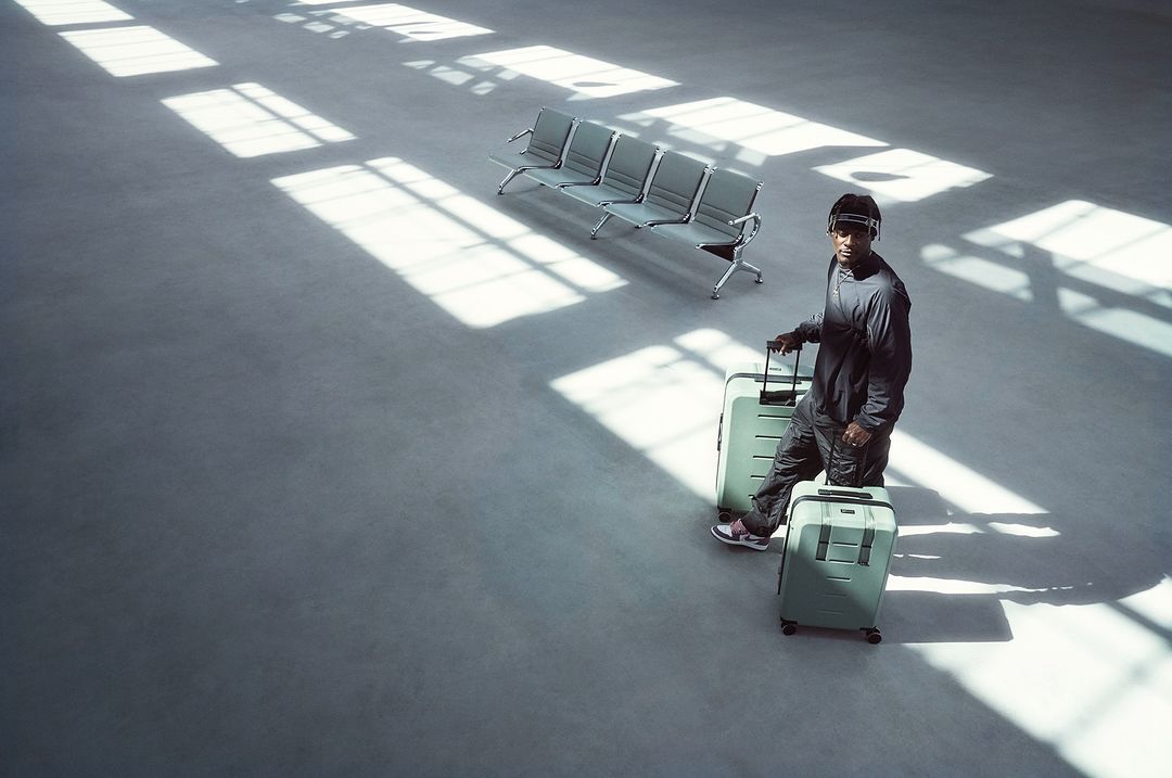 Adult male stands with two light green suitcases in a bright airport terminal with empty seats and natural light from overhead windows.