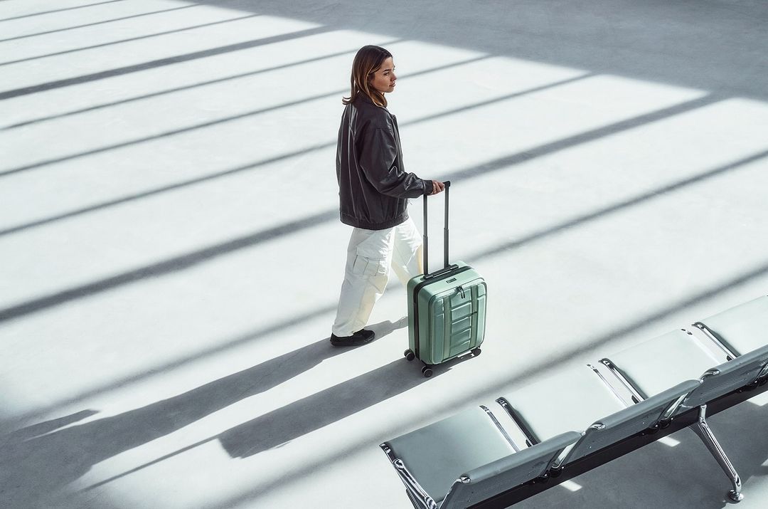 Woman with a rolling suitcase walks in an airy indoor space featuring white floors and rows of metal and white plastic seating.