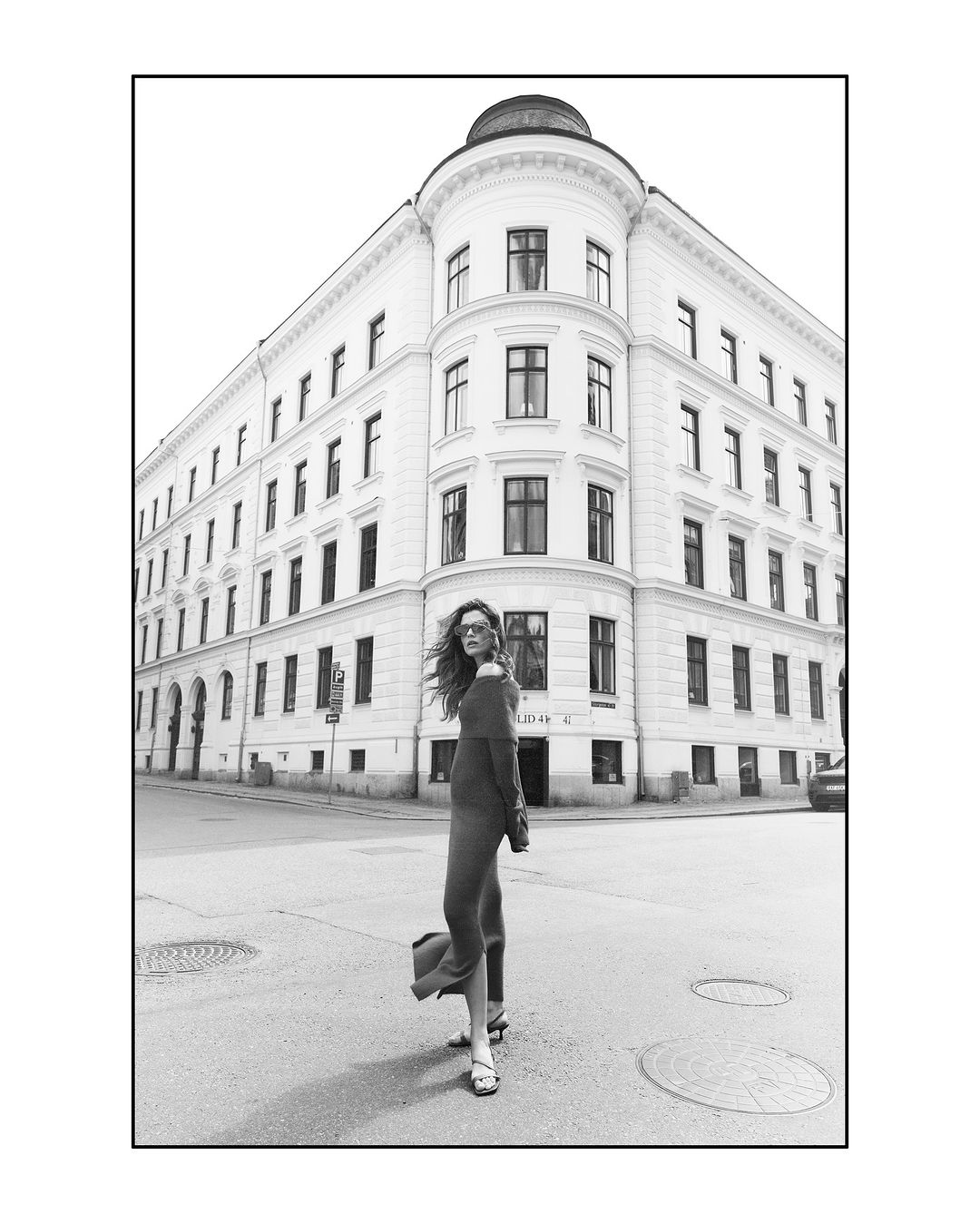 Model standing on a city street corner in front of a classical European building, captured in monochrome.