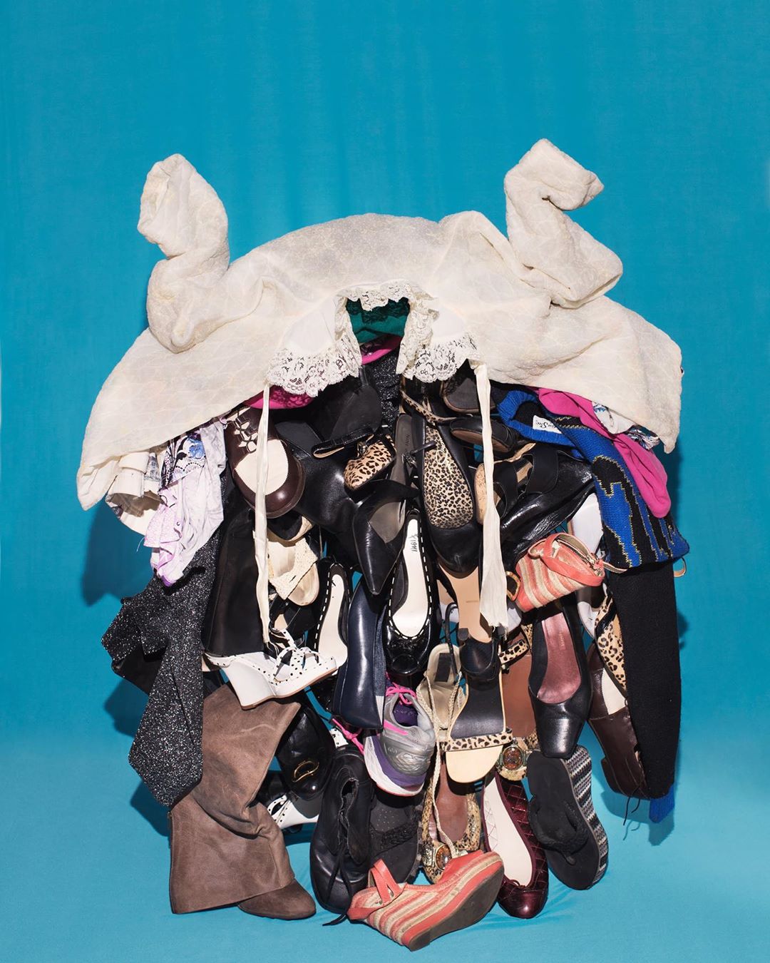 Pile of assorted women's shoes and clothing, including heels, flats, boots, and a white lacy jacket against a blue background.