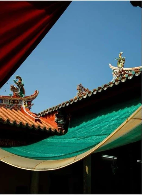 Eye-level view of an ornate Asian temple roof with colorful carvings and tiled shingles, with partial views of blue sky and awnings.