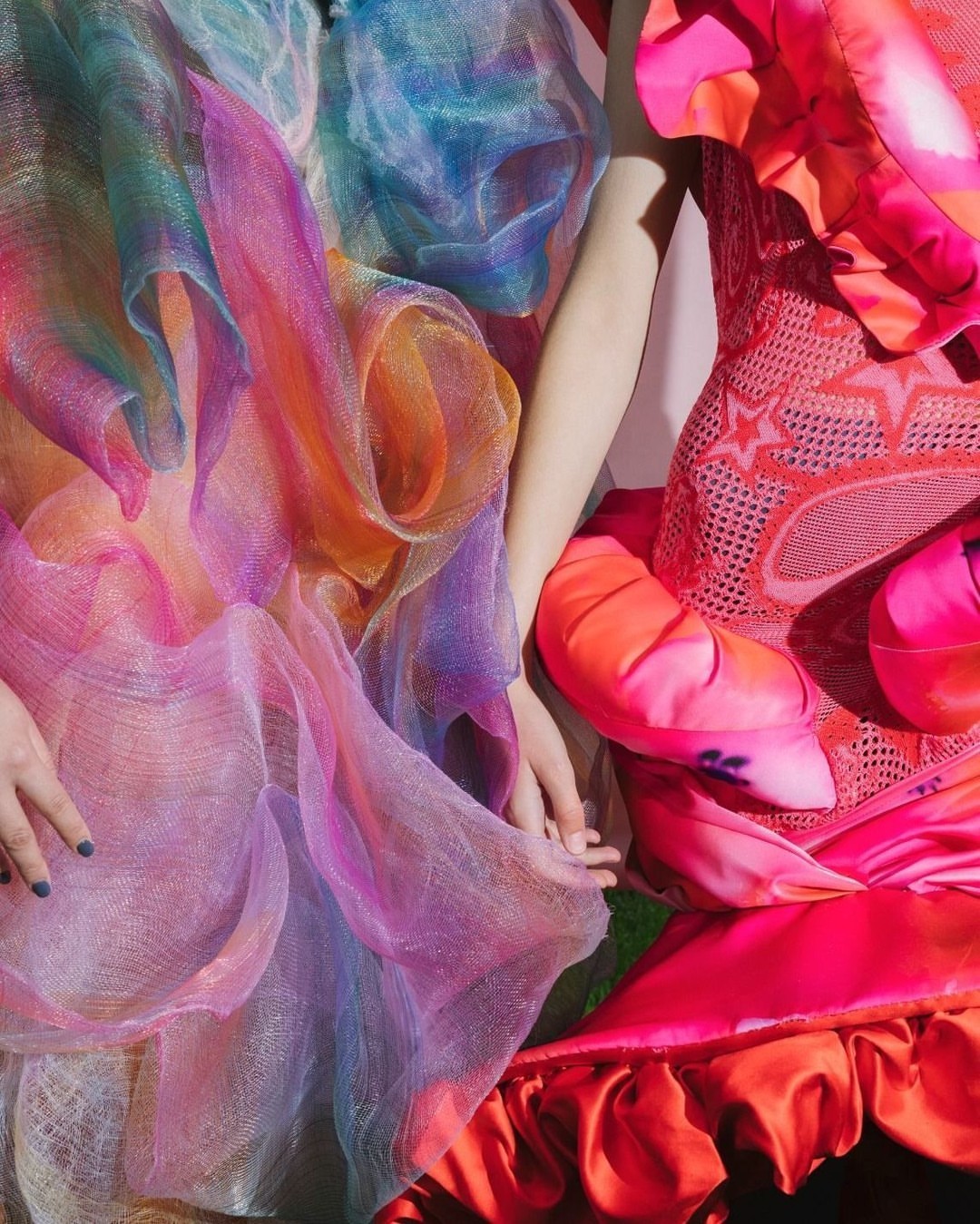 Two women hold hands wearing ornate and colorful tulle dresses in pink, red, orange, yellow, blue, and white, shot with bright, direct light.