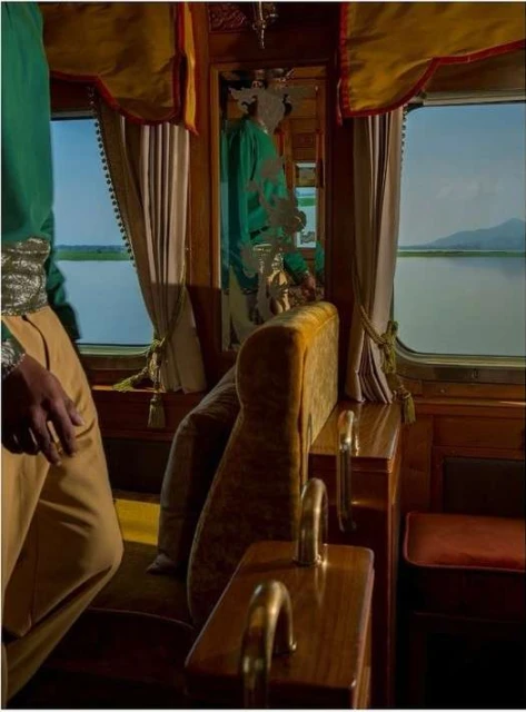 Interior view of a train car with a person partially visible in elaborate green and beige clothing, passing a lake landscape through the window.