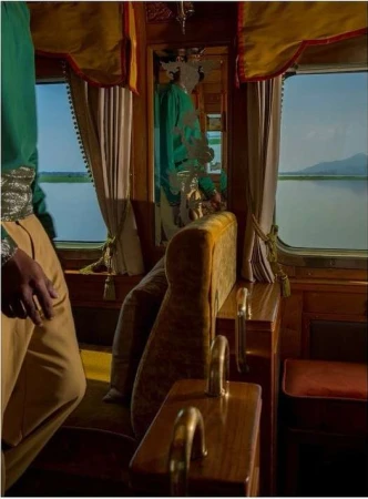 Interior view of a train car with a person partially visible in elaborate green and beige clothing, passing a lake landscape through the window.