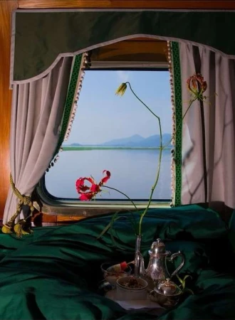 Still life of exotic flowers and antique silver tea service on emerald fabric inside a train car with a lake and mountains view outside.