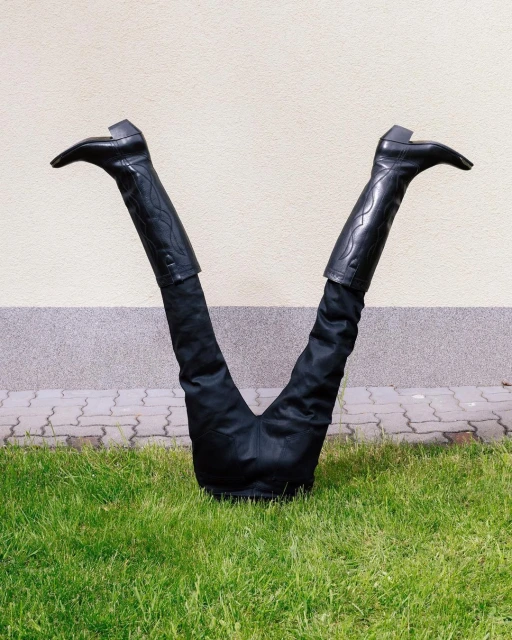 Upside-down person in black pants and leather boots on green grass with light-colored wall background outdoors on a sunny day.