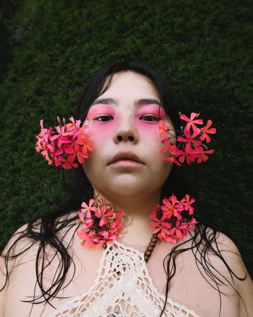 Portrait of a young woman with pink eye makeup and pink flowers around her face and shoulders, lying on a background of green leaves.