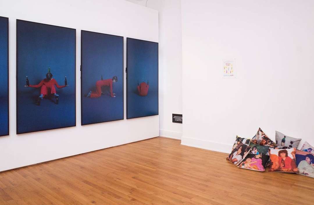 Four blue portrait paintings of a person in red clothing, displayed on a white gallery wall, with art-covered throw pillows on the floor.