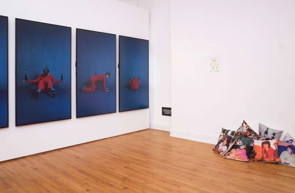 Four blue portrait paintings of a person in red clothing, displayed on a white gallery wall, with art-covered throw pillows on the floor.
