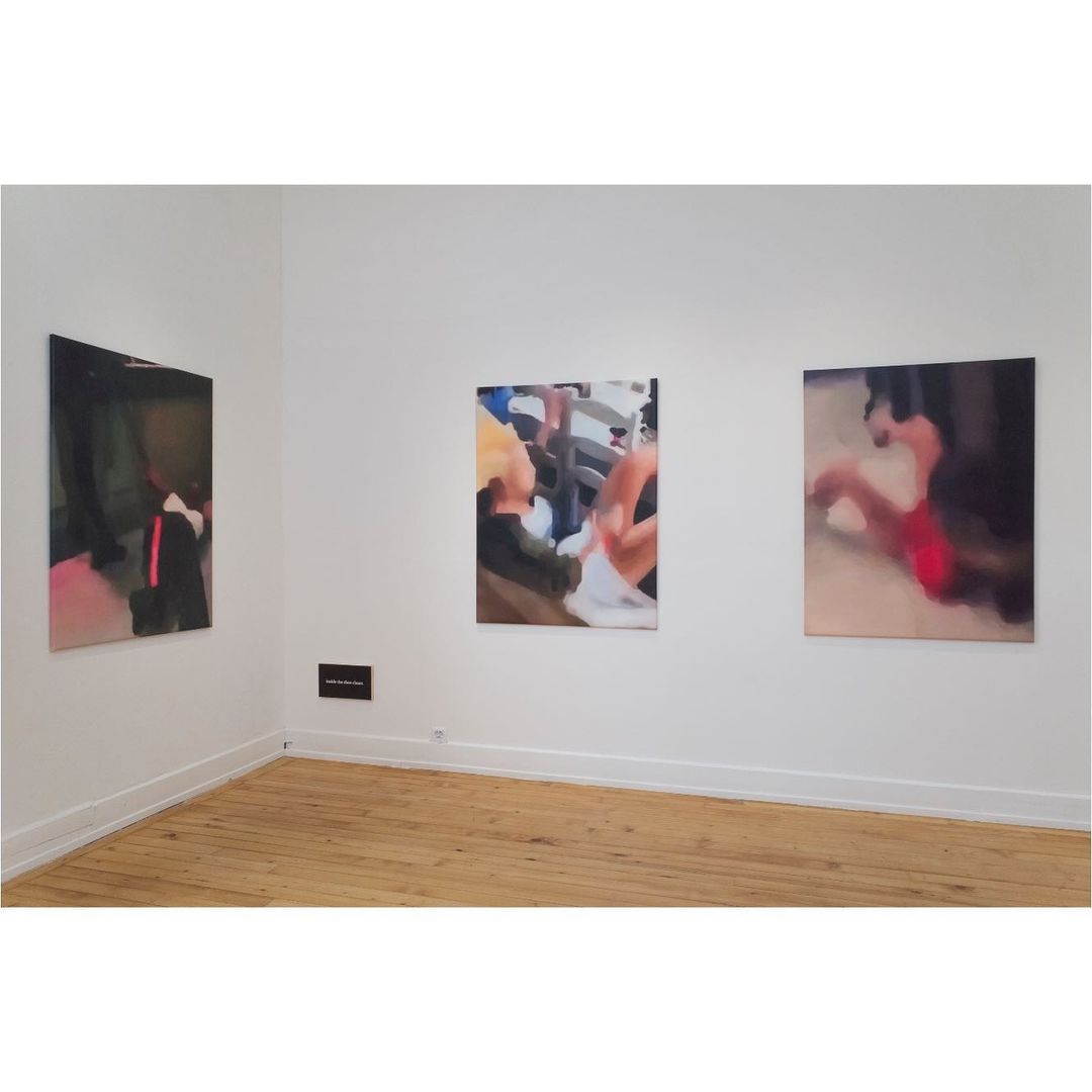 Art gallery view of three vibrant paintings hanging on a white wall, displaying figures and abstract forms in a brightly lit space with wood flooring.