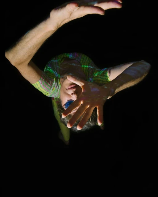 Adult with hands raised in a gesture illuminated by colorful patterned light against a dark backdrop in a low-angle portrait orientation.