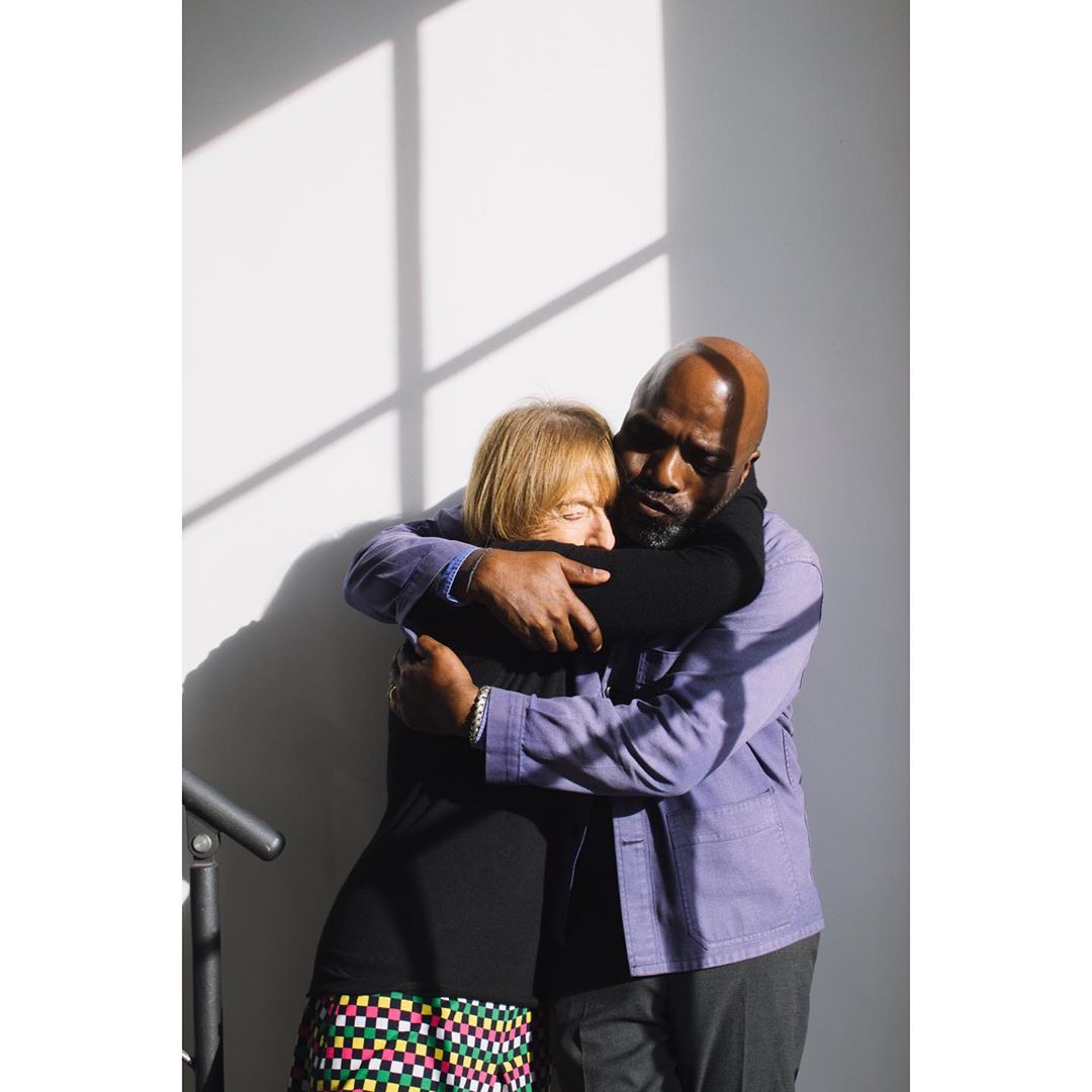 A couple embraces in a sunlit indoor space, featuring a man in a lavender jacket and a woman in a colorful skirt.
