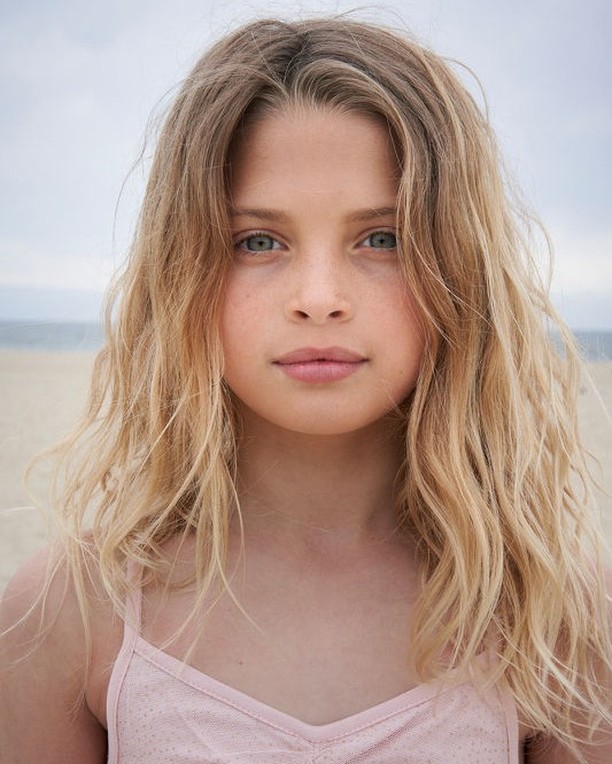 Close-up portrait of a young girl with blonde wavy hair and fair skin, looking at the camera on a sandy beach with an overcast sky.
