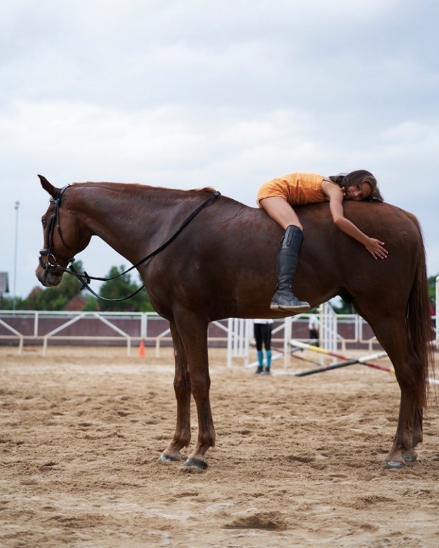 A woman is lying on a brown horse in an outdoor arena on a cloudy day. The woman wears a short, casual yellow dress and black riding boots.