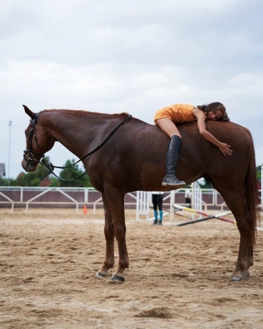 A woman is lying on a brown horse in an outdoor arena on a cloudy day. The woman wears a short, casual yellow dress and black riding boots.