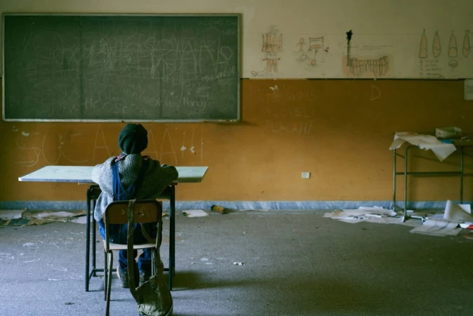 Child seated at a desk in an abandoned school classroom with graffiti on the blackboard and deteriorated conditions visible throughout.