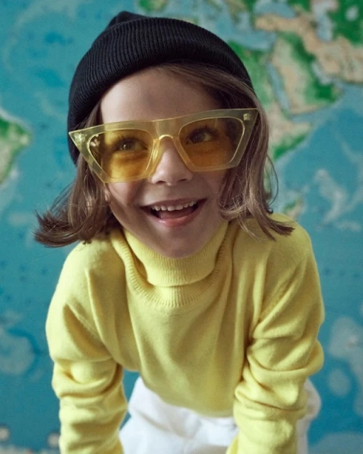A smiling child wearing yellow sunglasses and a black beanie stands in front of a blue and green world map.