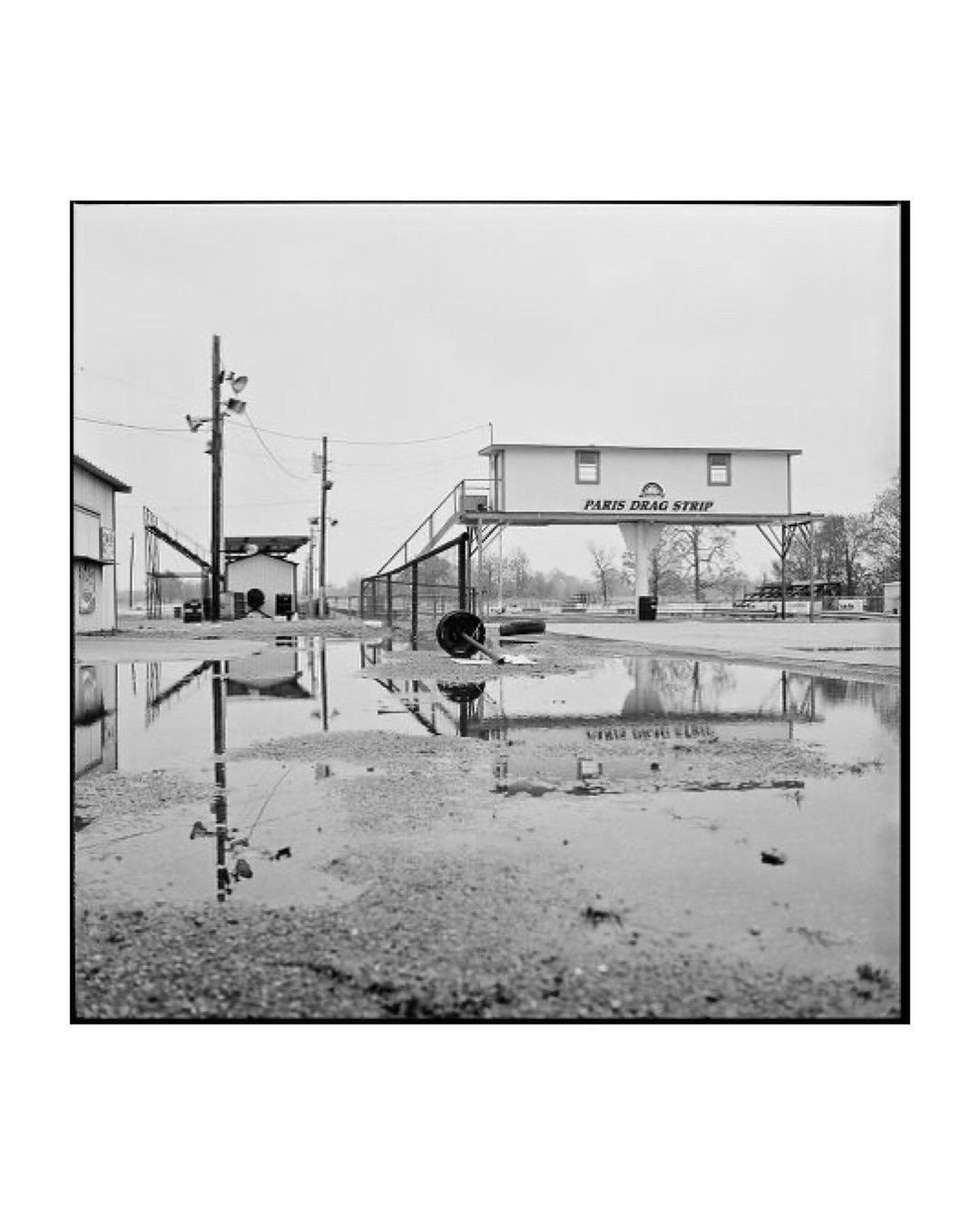 Black and white architectural shot of Paris Drag Strip building with water reflections on the ground and overcast sky.