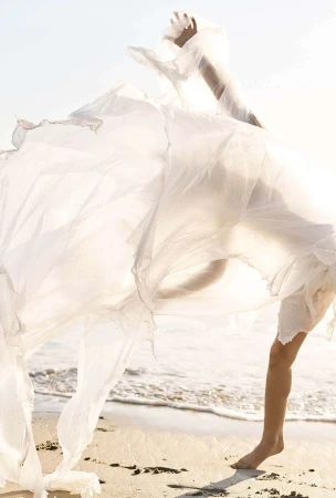 Model in flowing sheer white dress dancing on a sandy beach with ocean waves in the background.