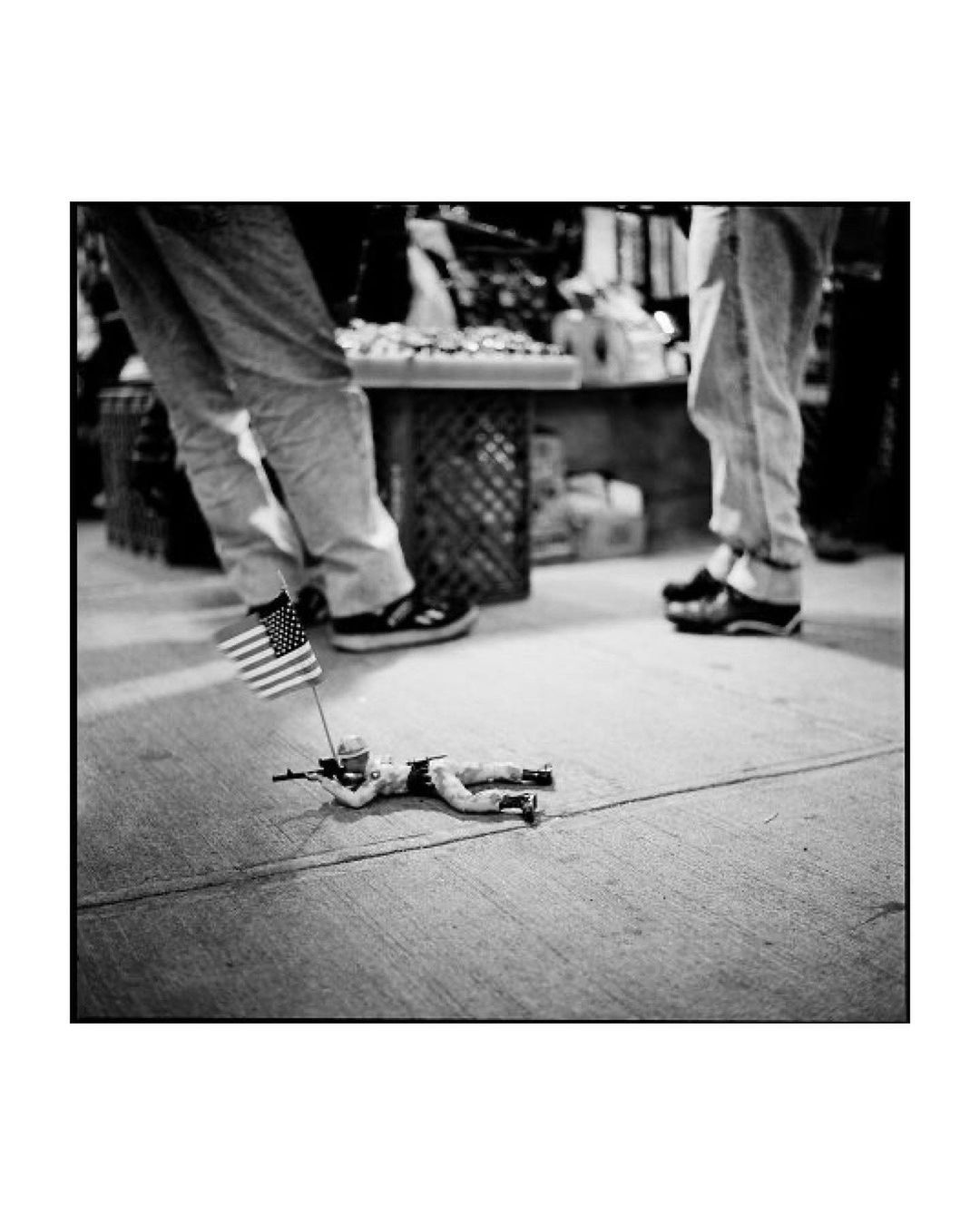 Black and white street photography of a toy soldier lying on the sidewalk with an American flag, near the feet of onlookers.