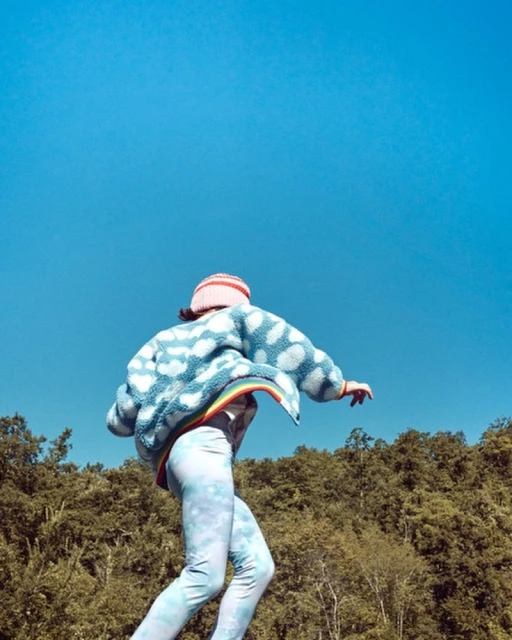 A person in a cloud-patterned jacket poses outdoors against a bright blue sky and a green tree line, captured in a sunny, medium shot.